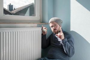 Man in warm clothes sits near the radiator and turns the thermostat to the minimum, making a warning gesture. Concept of increasing fuel costs. photo