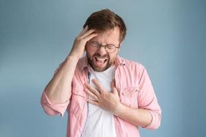 el hombre está experimentando un fuerte dolor de cabeza. sufre de migraña, se lleva una mano a la frente, la otra al corazón y grita de dolor. foto