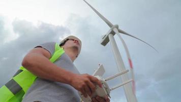 homem engenheiro de ecologia profissional de uniforme e capacete segurando o controle remoto para voar drone trabalhando no fundo dos moinhos de vento. serviço de parques eólicos. alternativa à energia elétrica. video