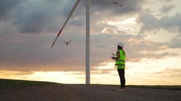 Professional Ecology Engineer in uniform and helmet holding remote controller for flying drone working at wind turbine on beautiful sunset background. Alternative to electrical energy.