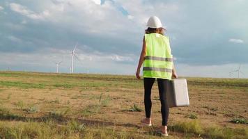 Woman Professional Ecology Engineer in uniform and helmet with special equipment in hand goes to service a windmill on beautiful sky and field background. Alternative energy concept. Slow motion. video