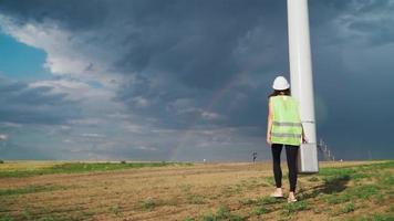 Woman Professional Ecology Engineer in uniform and helmet with special equipment in hand goes to service a windmill on beautiful sky and field background. Alternative energy concept. Slow motion.