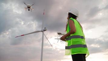 Professional Ecology Engineer in uniform and helmet holding remote controller for flying drone working at wind turbine on beautiful sunset background. Alternative to electrical energy. video