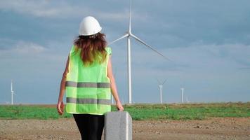 Woman Professional Ecology Engineer in uniform and helmet with special equipment in hand goes to service a windmill on beautiful sky and field background. Alternative energy concept. Slow motion. video