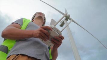 Mens professioneel ecologie ingenieur in uniform en helm Holding afgelegen controleur voor vliegend dar werken Aan achtergrond van windmolens. wind boerderij onderhoud. alternatief naar elektrisch energie. video