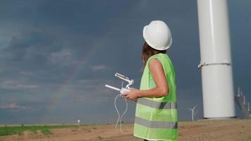 Professional Ecology Engineer in uniform and helmet holding remote controller for flying drone working at wind turbine on beautiful sunset background. Alternative to electrical energy. video