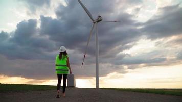 femme ingénieure en écologie professionnelle en uniforme et casque avec équipement spécial à la main va entretenir un moulin à vent sur fond de ciel et de champ magnifique. concept d'énergie alternative. ralenti.