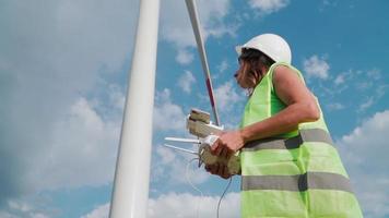 Professional Ecology Engineer in uniform and helmet holding remote controller for flying drone working on background of windmills. Wind farm service. Alternative to electrical energy. video