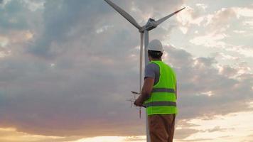 Man Professional Ecology Engineer in uniform and helmet holding remote controller for flying drone working at wind turbine on beautiful sunset background. Alternative to electrical energy. video