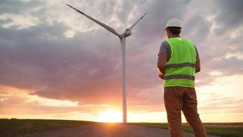 Man Professional Ecology Engineer in uniform and helmet holding remote controller for flying drone working at wind turbine on beautiful sunset background. Alternative to electrical energy. video