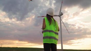 Professional Ecology Engineer in uniform and helmet holding remote controller for flying drone working at wind turbine on beautiful sunset background. Alternative to electrical energy. video