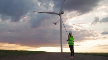 professionell ekologi ingenjör i enhetlig och hjälm innehav avlägsen kontrollant för flygande Drönare arbetssätt på vind turbin på skön solnedgång bakgrund. alternativ till elektrisk energi. video