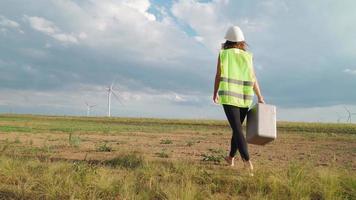 kvinna professionell ekologi ingenjör i enhetlig och hjälm med särskild Utrustning i hand går till service en väderkvarn på skön himmel och fält bakgrund. alternativ energi begrepp. långsam rörelse. video