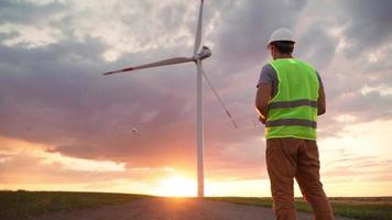 uomo professionale ecologia ingegnere nel uniforme e casco Tenere a distanza controllore per volante fuco Lavorando a vento turbina su bellissimo tramonto sfondo. alternativa per elettrico energia. video