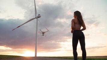 Sexy slim woman in sportswear with a remote control in her hands lifts the drone up against the backdrop of a beautiful sunset and windmills. Slow motion. video