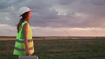 vrouw professioneel ecologie ingenieur in uniform en helm met speciaal uitrusting in hand- gaat naar onderhoud een windmolen Aan mooi zonsondergang achtergrond. alternatief energie concept. langzaam beweging. video