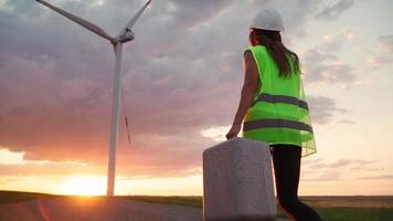 Woman Professional Ecology Engineer in uniform and helmet with special equipment in hand goes to service a windmill on beautiful sunset background. Alternative energy concept. Slow motion.