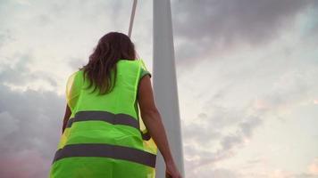 vrouw professioneel ecologie ingenieur in uniform en helm met speciaal uitrusting in hand- gaat naar onderhoud een windmolen Aan mooi lucht en veld- achtergrond. alternatief energie concept. langzaam beweging. video