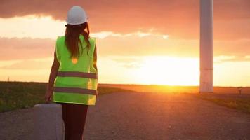 donna professionale ecologia ingegnere nel uniforme e casco con speciale attrezzatura nel mano va per servizio un' mulino a vento su bellissimo tramonto sfondo. alternativa energia concetto. lento movimento. video