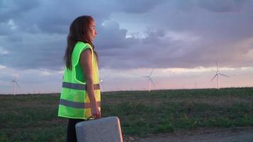 donna professionale ecologia ingegnere nel uniforme e casco con speciale attrezzatura nel mano va per servizio un' mulino a vento su bellissimo cielo e campo sfondo. alternativa energia concetto. lento movimento. video