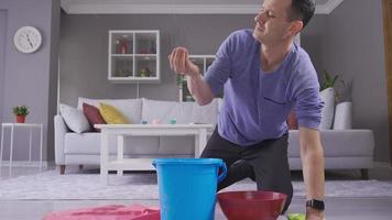 Sad man waiting for roof repair service looking at water falling from ceiling into plastic buckets. Man holding bucket under leaky ceiling and mopping floors. video