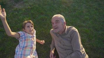 The boy asks his grandfather a question. The old man and his grandson lying on the grass. video