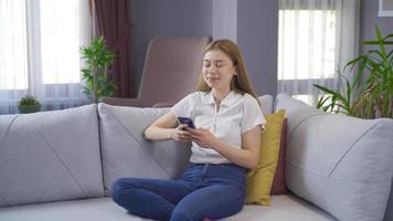 sorridente giovane ragazza sms su il Telefono a casa. ragazza seduta su divano a casa sms su sua Telefono e sorridente. video