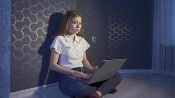 la mujer joven que trabaja con una computadora portátil por la noche está sola. mujer joven sentada en el piso en casa trabajando con una computadora portátil. video