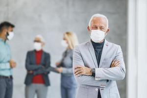 un hombre de negocios con máscara protectora de pie cruzado de brazos en la oficina durante la pandemia de covid-19 y mirando la cámara foto