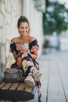 mujer usando un teléfono inteligente y navegando por la red mientras se sienta en un banco en la ciudad mediterránea foto