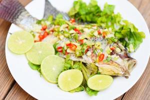 Pescado de tilapia al vapor con salsa de chile y lima limón con hierbas y verduras en el plato, comida tilapia cocida pescado lima al vapor - comida tailandesa foto