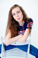 portrait of a young and serious girl sitting on a chair, on a white background, with long hair, wearing a tight T-shirt and jeans photo
