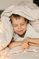portrait of a smiling boy lying under a white blanket and with a garland. child's christmas morning. Boy wake up in the morning photo