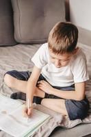 niño hace la tarea mientras está sentado en el sofá gris. un adolescente caucásico escribe en un libro de trabajo en el sofá. educar en casa a un niño de 9 años. aprendizaje libre foto