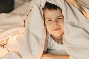 portrait of a smiling boy lying under a white blanket and with a garland. child's christmas morning. Boy wake up in the morning, copy space photo