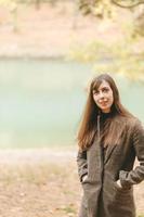 retrato de una chica con el pelo largo y oscuro, con un abrigo marrón, en el fondo de un lago otoñal. marco vertical de una chica en la calle en otoño, espacio de copia foto