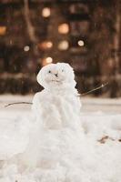 primer plano de un pequeño muñeco de nieve en invierno, en el contexto de las luces nocturnas. diversión de invierno durante las nevadas. nieve pegajosa hombre hecho de nieve foto