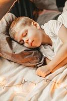 portrait of a boy sleeping under a white blanket and with a garland. christmas night baby. Little boy sleeping on gray pillow, vertical frame photo