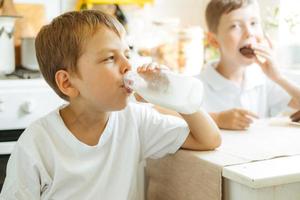 A boy is drinking milk from a bottle in the kitchen at home. Morning breakfast with milk. Happy child in a white T-shirt drinks milk from a bottle photo