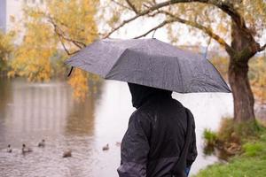 un hombre con una chaqueta con capucha, de espaldas con un paraguas en las manos, contra el fondo del lago
