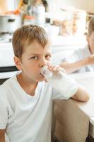 A boy is drinking milk from a bottle in the kitchen at home. Morning breakfast with milk. Happy child in a white T-shirt drinks milk from a bottle photo