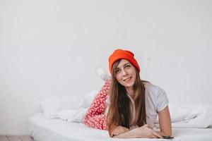 chica graciosa sonriente con sombrero rojo y pijama de navidad, yace en la cama con sábanas blancas. vacaciones para navidad. buen humor navideño chica. chica con camiseta blanca y pijama foto