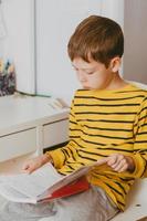 boy doing homework at home at the table. Schoolboy reads a book at home. Boy's room. Red-haired boy in a striped yellow long-sleeved t-shirt photo