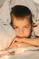 portrait of a serious boy lying under a white blanket and with a garland. child's christmas morning. Boy wake up in the morning, vertical frame photo