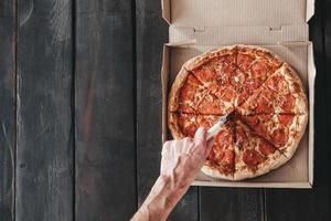 la mano masculina corta la pizza de pepperoni con un cuchillo redondo sobre un fondo de madera oscura. pizza deliciosa y fresca en una caja con trozos redondos de salchicha, sobre un fondo de madera. delivery de pizza foto