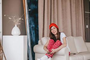 smiling girl in a red hat and Christmas pajamas sitting on the couch. holidays for christmas. Good Christmas mood girl. photo