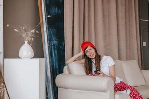 smiling girl in a red hat and Christmas pajamas sitting on the couch. holidays for christmas. Good Christmas mood girl. photo