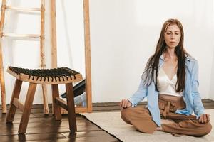 girl with long and dark hair sitting lotus pose meditating on floor light room interior. Yoga practice