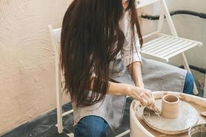 A girl with long hair sculpts a clay pot on a potter's wheel from a special wooden tool. Creative workshop. The women sculpts from clay. Working atmosphere in the workshop photo