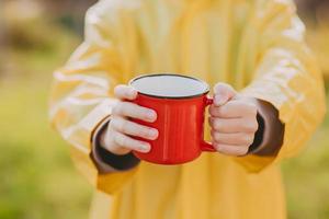 las manos de un niño con un impermeable amarillo sostienen chocolate caliente en una taza roja. foto acogedora con una taza. concepto de campamento. foto de otoño con una taza de cacao en la calle